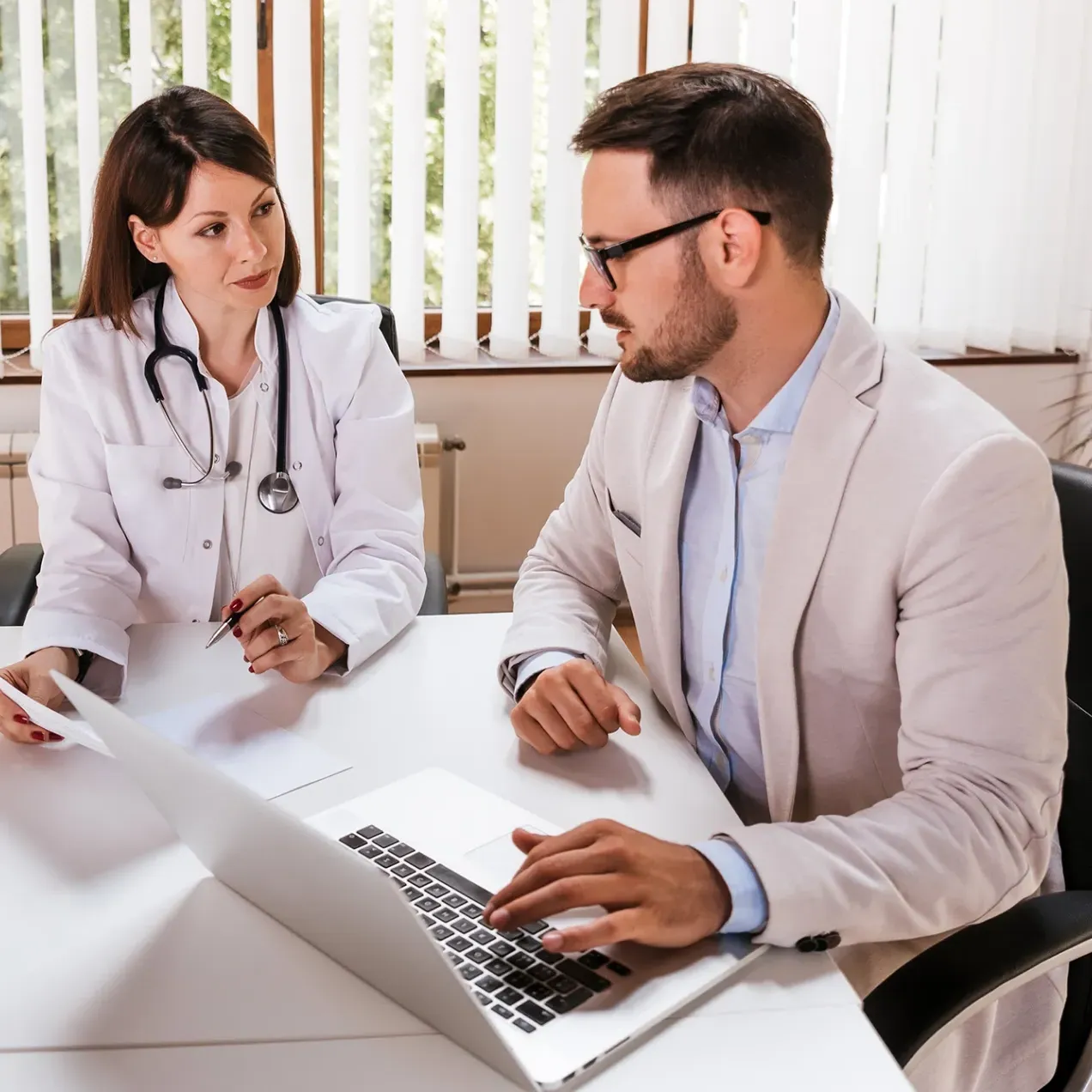 Doctor in a white coat with a stethoscope having a discussion with a professional man using a laptop in a bright office setting.